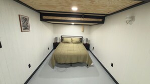 Une chambre à coucher avec un lit placé au centre d’un plancher blanc, flanqué de tables de chevet avec des lampes, un plafond en bois et un tableau encadré au mur.