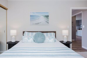 A bedroom with a bed, two bedside tables with lamps, a wall-mounted picture of a beach, and a view into the adjacent kitchen.