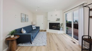 A modern living room with a blue sofa, a fireplace, and a view of a wooden deck through large sliding glass doors. - The Coastal Retreat, Scenic Harbor, Ocean Views, Walk to Beach, Restaurants (El Granada)