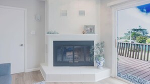 A modern living room with a fireplace, a white door, and a view of a wooden deck. - The Coastal Retreat, Scenic Harbor, Ocean Views, Walk to Beach, Restaurants (El Granada)