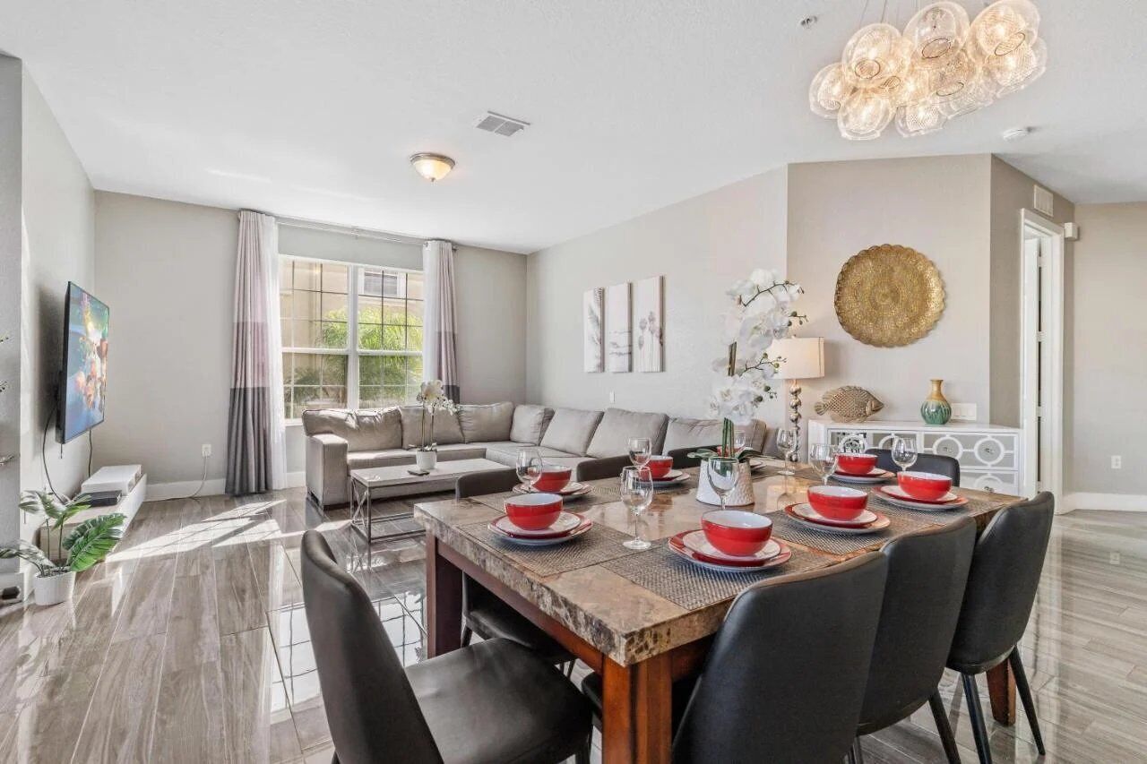 A modern living room with a dining area, featuring a large window, a sofa, a TV, and a dining table set with red and white plates.