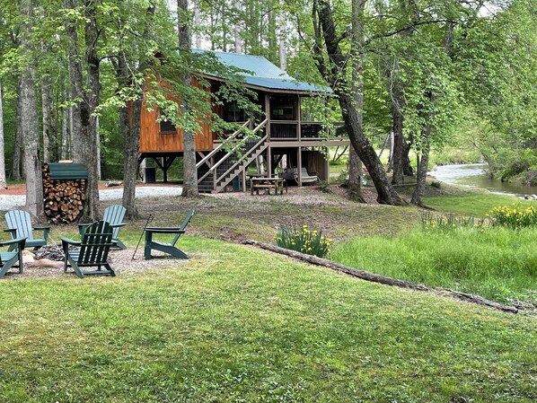 A cabin with a deck, surrounded by trees and a grassy area with outdoor seating.