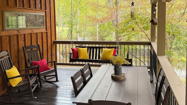 A wooden deck with a swing chair and a table, surrounded by a railing and a view of trees. - Your Perfect Creekside Hideaway (Rabun Gap)