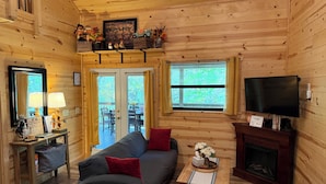 A cozy cabin interior with a sofa, television, fireplace, and a dining area visible through a door. - Your Perfect Creekside Hideaway (Rabun Gap)