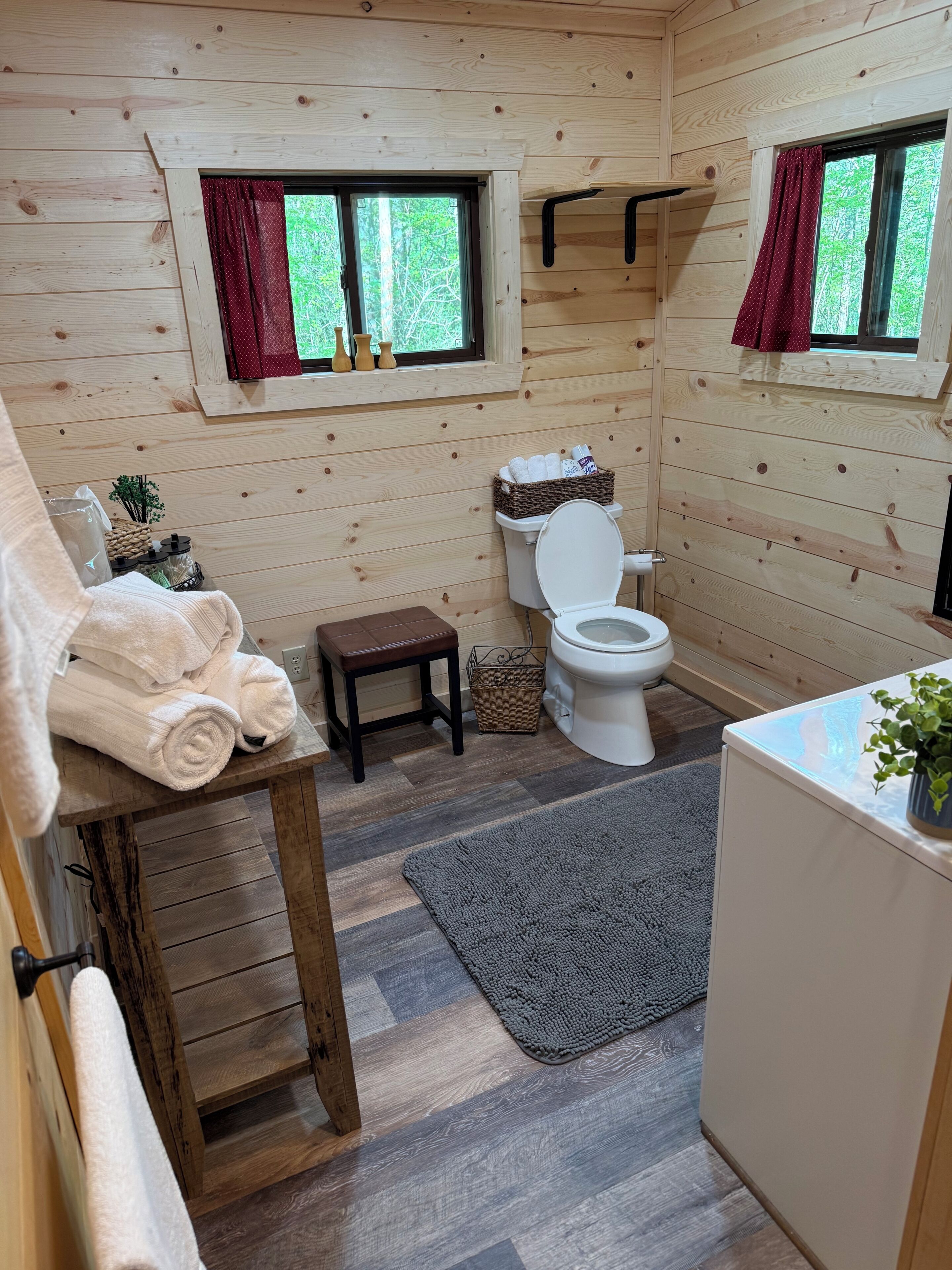 Une salle de bain rustique, aménagée dans une cabane en bois, avec des toilettes, une petite table sur laquelle sont posées des serviettes, une fenêtre avec des rideaux et un plancher en bois.