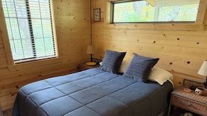 A wooden cabin bedroom with a large bed, two pillows, a nightstand with a lamp, and a window with blinds. - Your Perfect Creekside Hideaway (Rabun Gap)