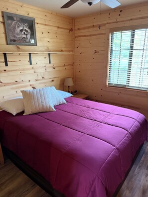 A wooden cabin room with a bed, a ceiling fan, a window with blinds, and a raccoon picture on the wall.