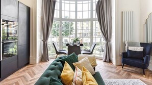 A room with a curved bay window, a dark green sofa with multiple cushions, a dark blue armchair, and a black cabinet with glass doors. - Relaxing Seaside Retreats (Brighton and Hove)
