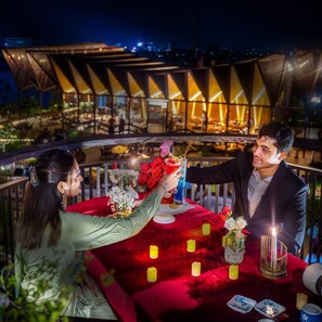 Una pareja brindando con bebidas en una mesa con mantel rojo, velas y vista a un edificio moderno al fondo.