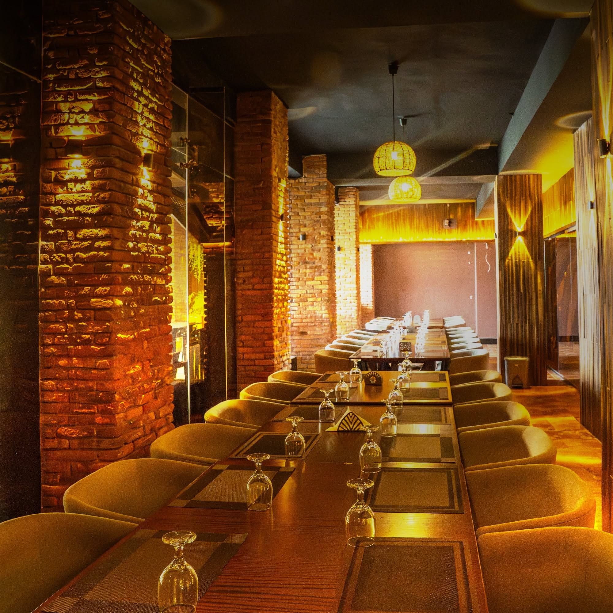 A restaurant with a long table set for dining, brick walls, and hanging pendant lights.