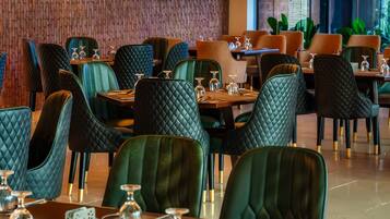 A restaurant with green upholstered chairs, wooden tables, and a brick wall in the background.