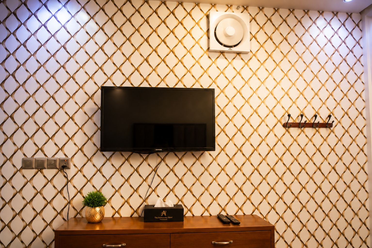 A hotel room with a flat-screen TV mounted on a patterned wall, a wooden dresser with a remote control, a plant, and a tissue box.