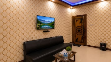 A modern living room with a black leather sofa, a glass coffee table with water bottles, a flat-screen TV on the wall, and a decorative door with a star and crescent symbol.