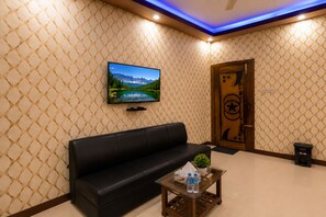 Una sala moderna con un sofá de cuero negro, una mesa de centro de vidrio con botellas de agua, un televisor de pantalla plana en la pared y una puerta decorativa con un símbolo de estrella y luna creciente.