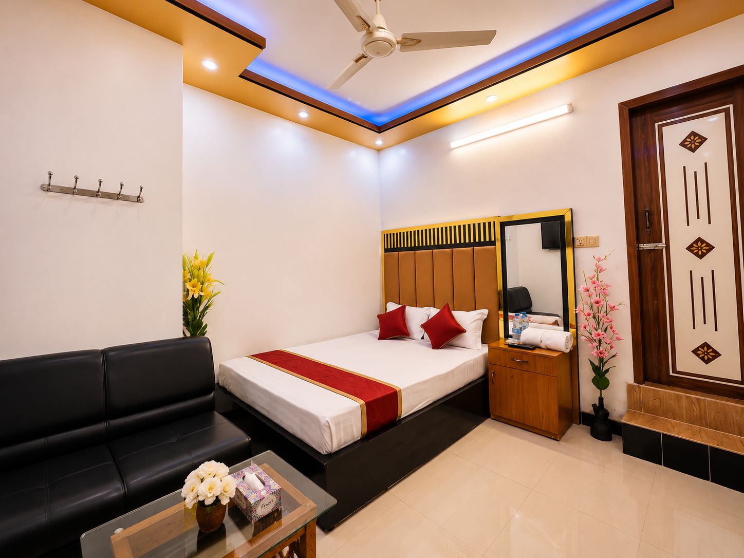 A hotel room with a bed, a sofa, a small table with a vase, a mirror, and a wooden door.