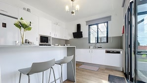 A modern kitchen with white cabinets, a microwave, a sink, and a window. There are two bar stools and a vase with flowers on the counter. - Villa Magn lia by Atlantic Holiday (Machico)
