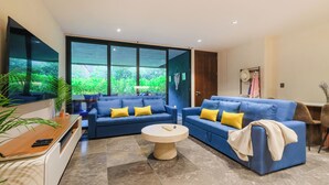 A modern living room with two blue sofas, a round coffee table, and a large window offering a view of greenery. - 2-bedroom condo in enchanting Tulum with AC (Tulum)