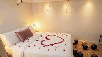 A bedroom with a bed, pillows, a heart-shaped arrangement of rose petals, balloons, and scattered rose petals on the floor.