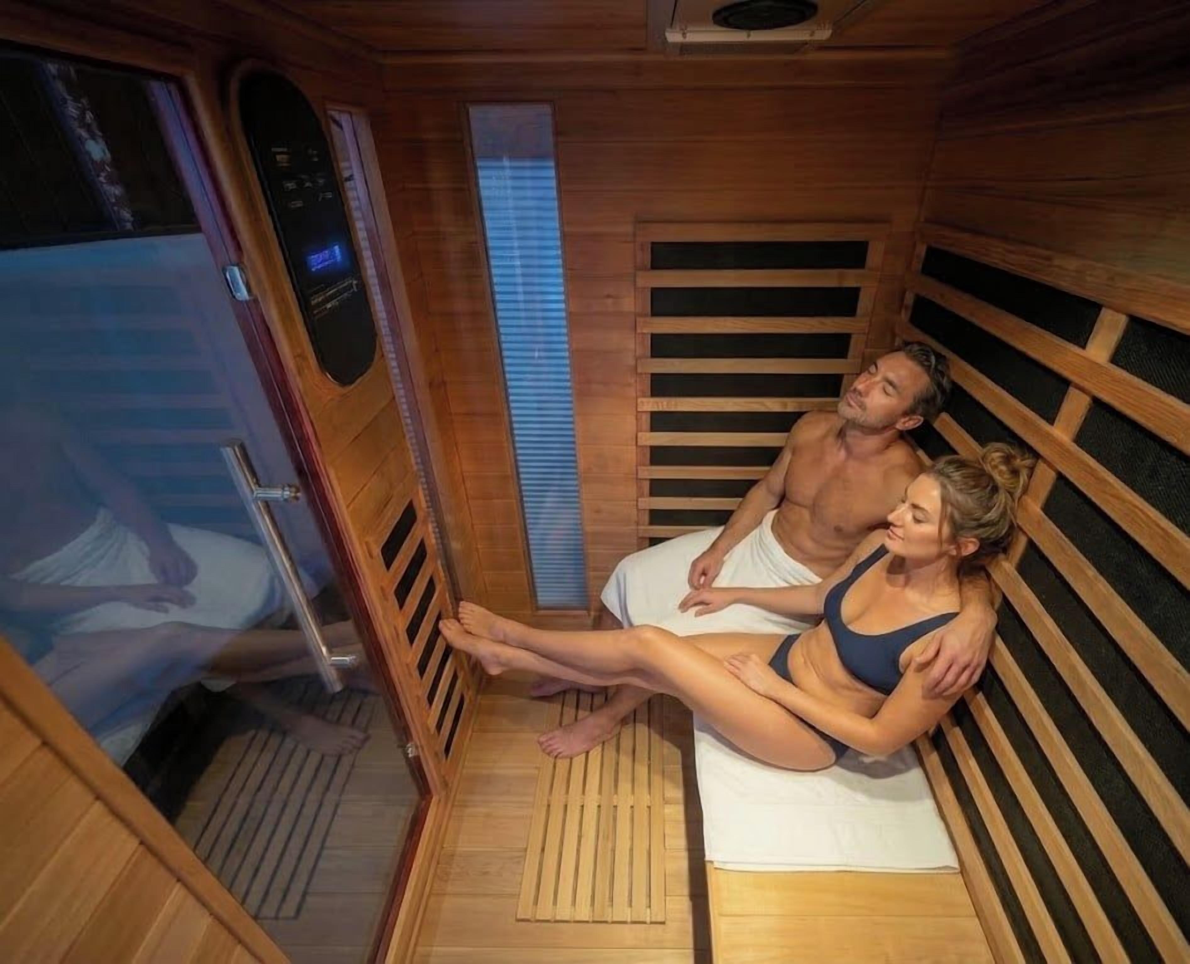 A sauna room with wooden walls and benches, a control panel, and a person lying on a bench.