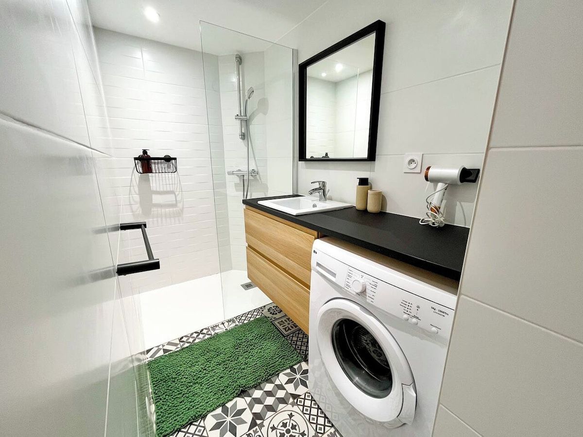 A modern bathroom with a glass shower enclosure, a washing machine, a sink, and a mirror.