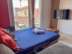 A hotel room with a bed, a TV, a small table, and a view of buildings outside.