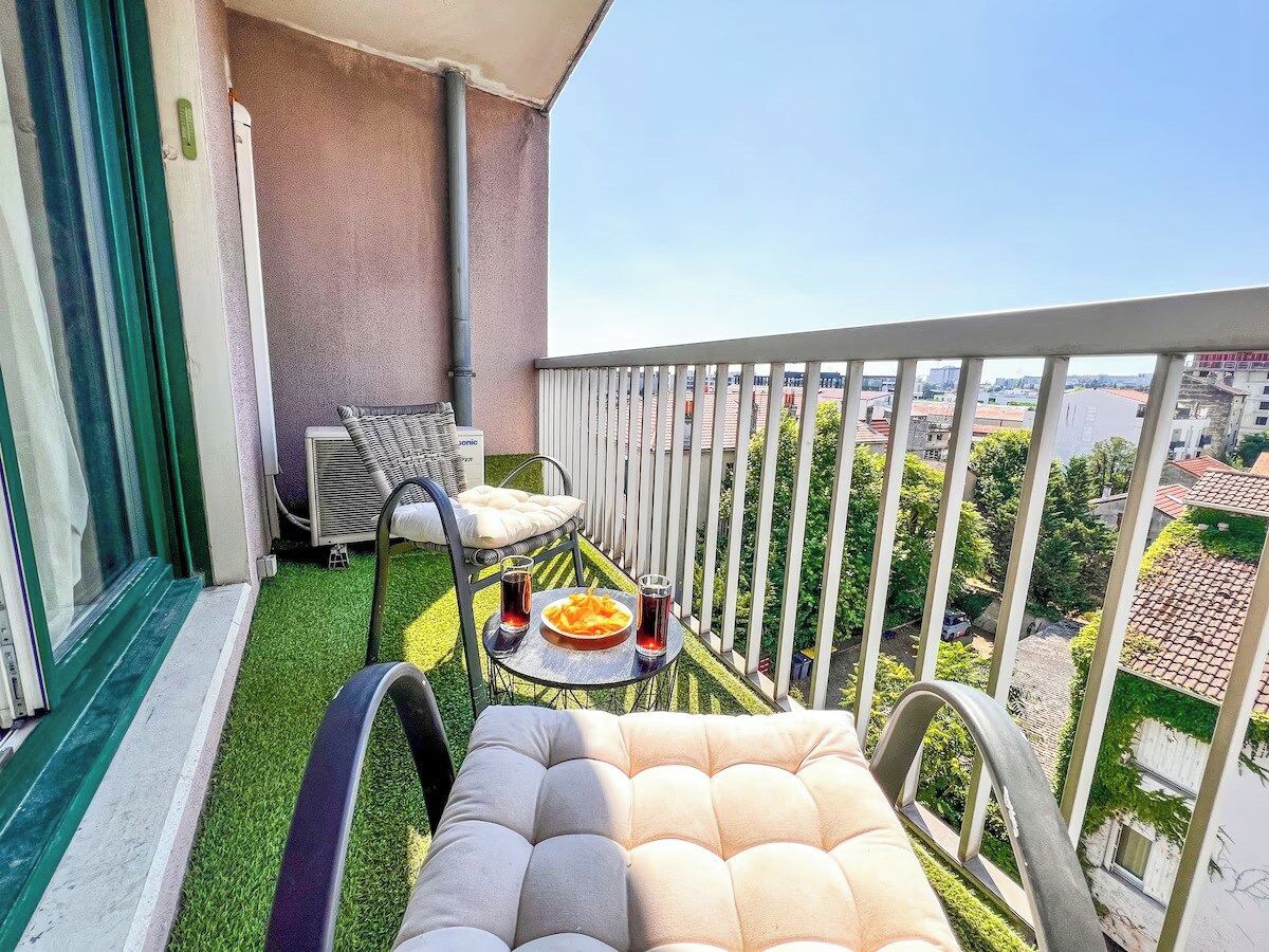 A balcony with a cushioned chair, a small table with a plate of food and drinks, and a view of the cityscape.
