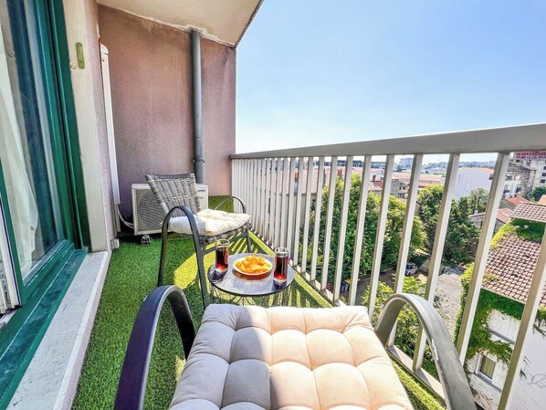 A balcony with a cushioned chair, a small table with a plate of food and drinks, and a view of the cityscape.