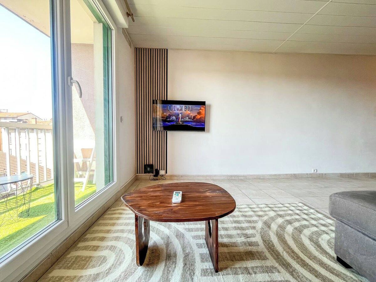 Ein modernes Wohnzimmer mit großem Fenster, einem an der Wand befestigten Flachbildfernseher, einem runden Holztisch und einem grauen Sofa.