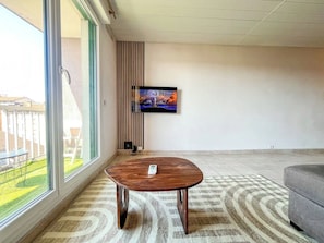 A modern living room with a large window, a flat-screen TV mounted on the wall, a round wooden coffee table, and a grey sofa.