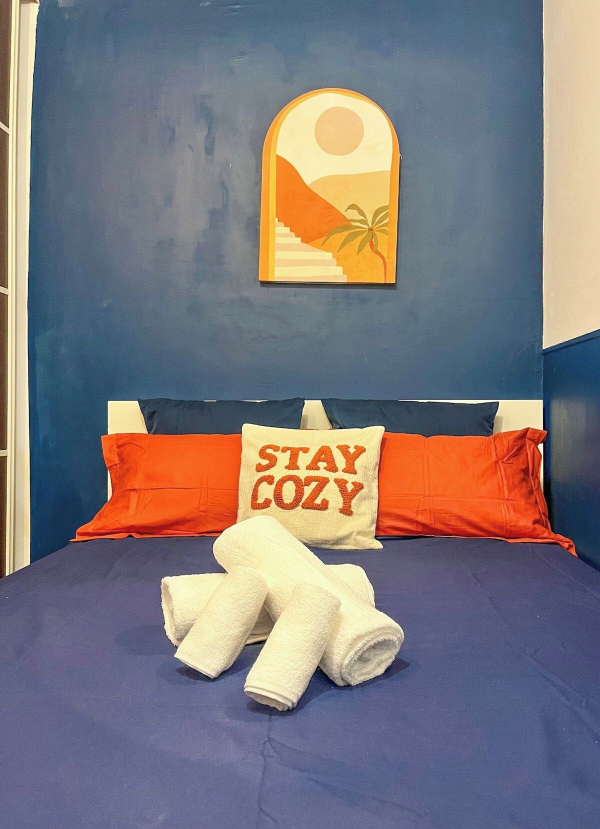 Un lit avec une tête de lit bleue, des oreillers orange et blancs, un coussin orné de l’inscription « RESTEZ AU CHAUD » et une serviette blanche.