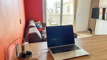 Una computadora portátil sobre una mesa de madera, con una regleta y cable, una pared roja, un balcón con sillas y vistas a edificios.