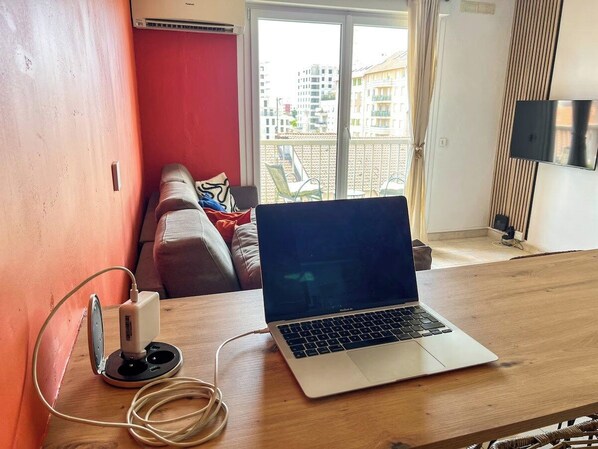 A laptop on a wooden table with a power strip and cable, a red wall, a balcony with chairs, and a view of buildings.