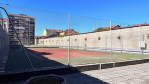 Ein eingezäunter Sportplatz mit Basketballkorb, umgeben von einem Maschendrahtzaun und Gebäuden im Hintergrund.