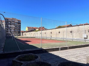 Een omheind sportterrein met een basketbalring, omgeven door een hek en gebouwen op de achtergrond.
