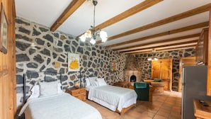Una habitación rústica con dos camas, una pared de piedra, vigas de madera, chimenea y una zona de cocina.