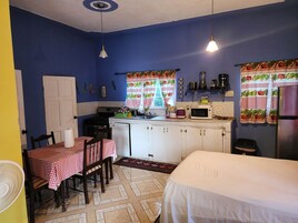 A compact kitchen with a stove, microwave, and sink. There is a dining table with chairs and a bed in the room.