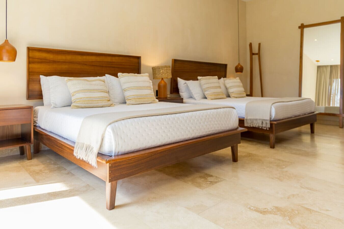 A hotel room with two wooden beds, each with white bedding and striped pillows. There are bedside tables with lamps and a large mirror on the wall.