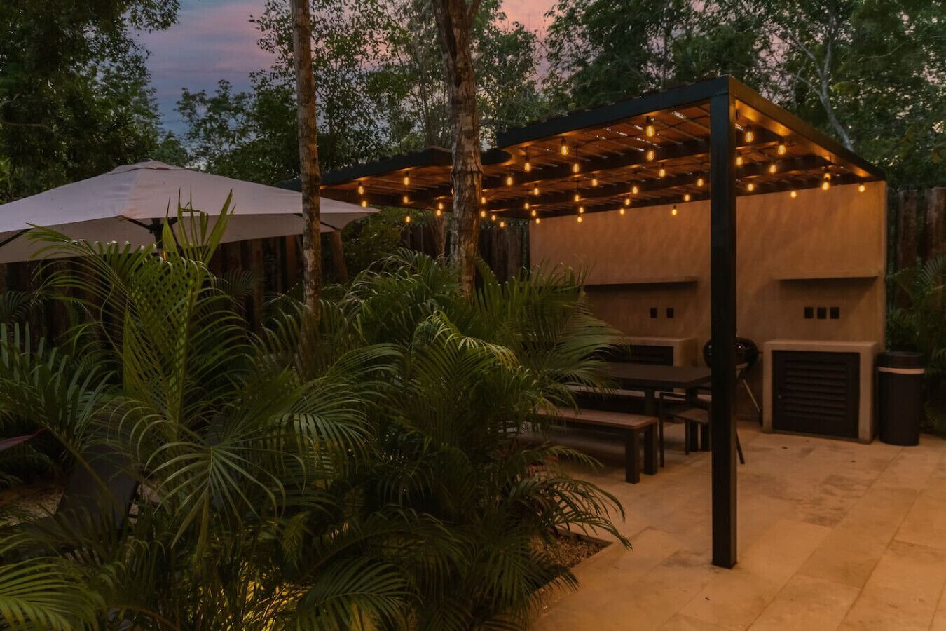 Un espace extérieur aménagé en terrasse, avec des tables et des bancs en bois, entouré d’une végétation luxuriante et illuminé de guirlandes lumineuses.
