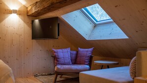 A cozy attic room with wooden walls, a large skylight, a comfortable chair with cushions, a small round table, and a flat-screen TV mounted on the wall.