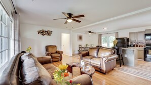 A spacious living room with leather sofas, a coffee table, and a ceiling fan. A kitchen with a refrigerator and stove is visible in the background. - Group Getaway w/ Fenced Yard & Pool Table in Wrens (Wrens)