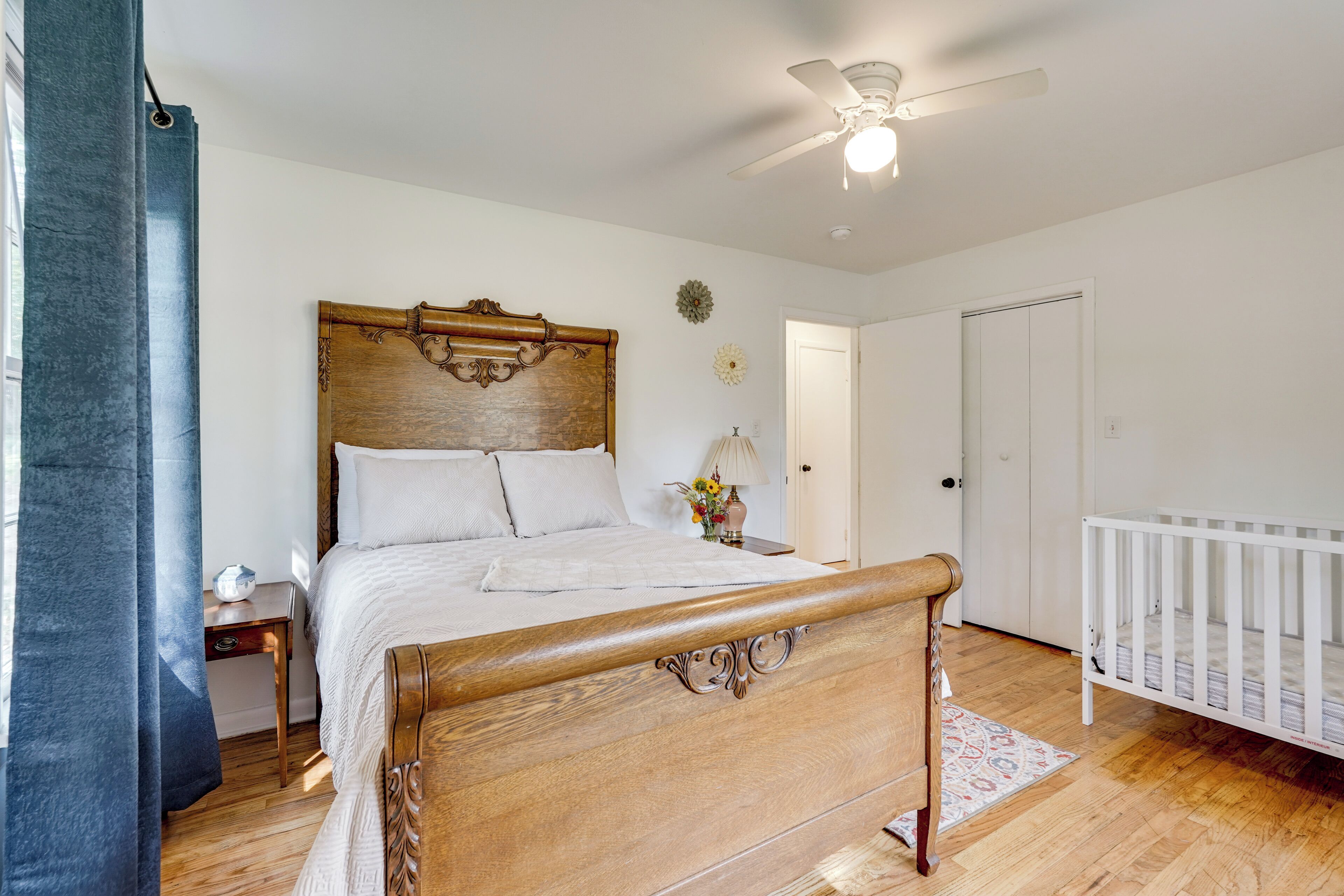 A bedroom with a wooden bed, a white crib, a ceiling fan, and a nightstand with a lamp.