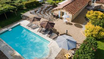 Une piscine entourée de chaises longues et de parasols, elle-même bordée d’un jardin et d’un bâtiment dont le toit est recouvert de tuiles.