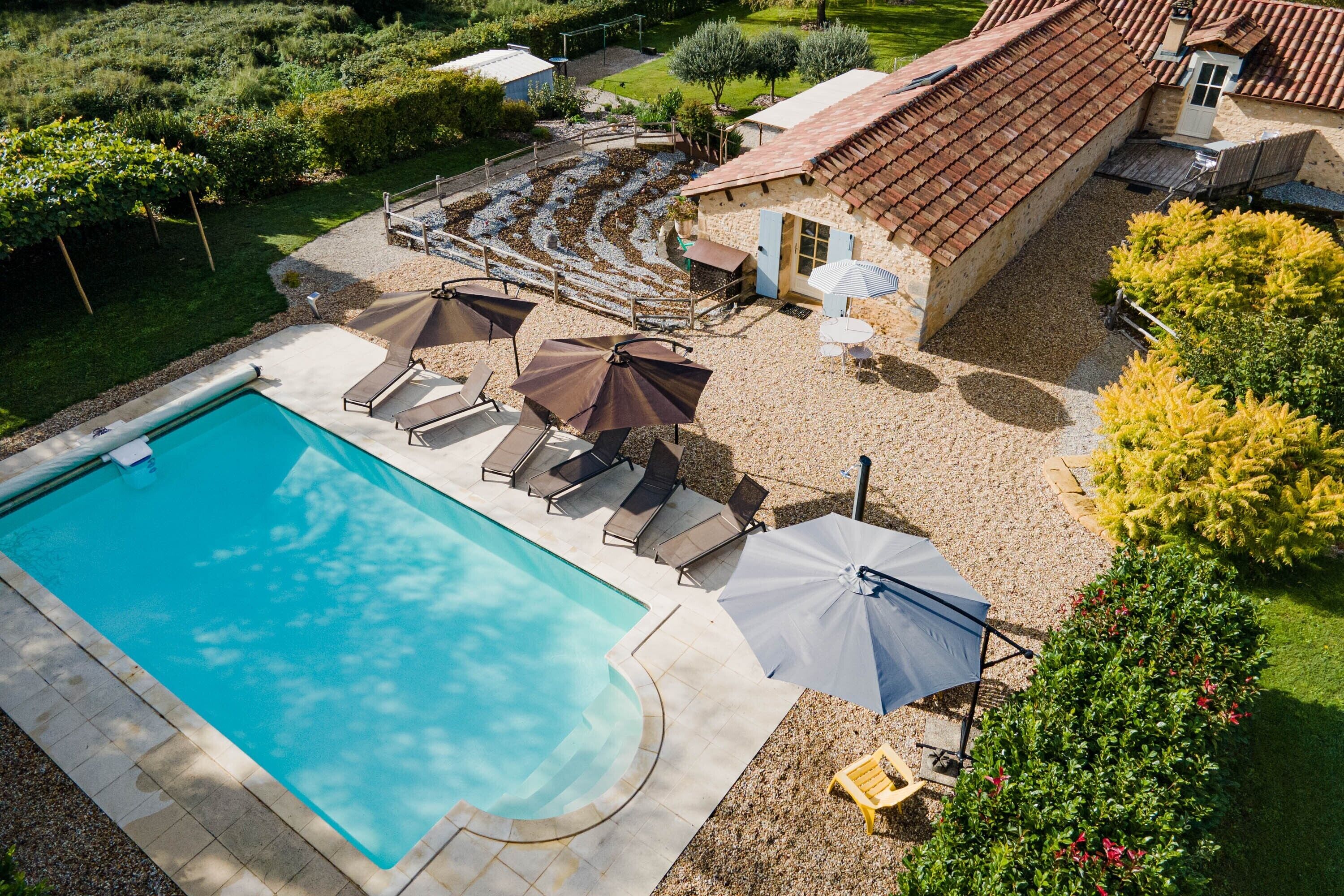A swimming pool with lounge chairs and umbrellas, surrounded by a garden and a building with a tiled roof.