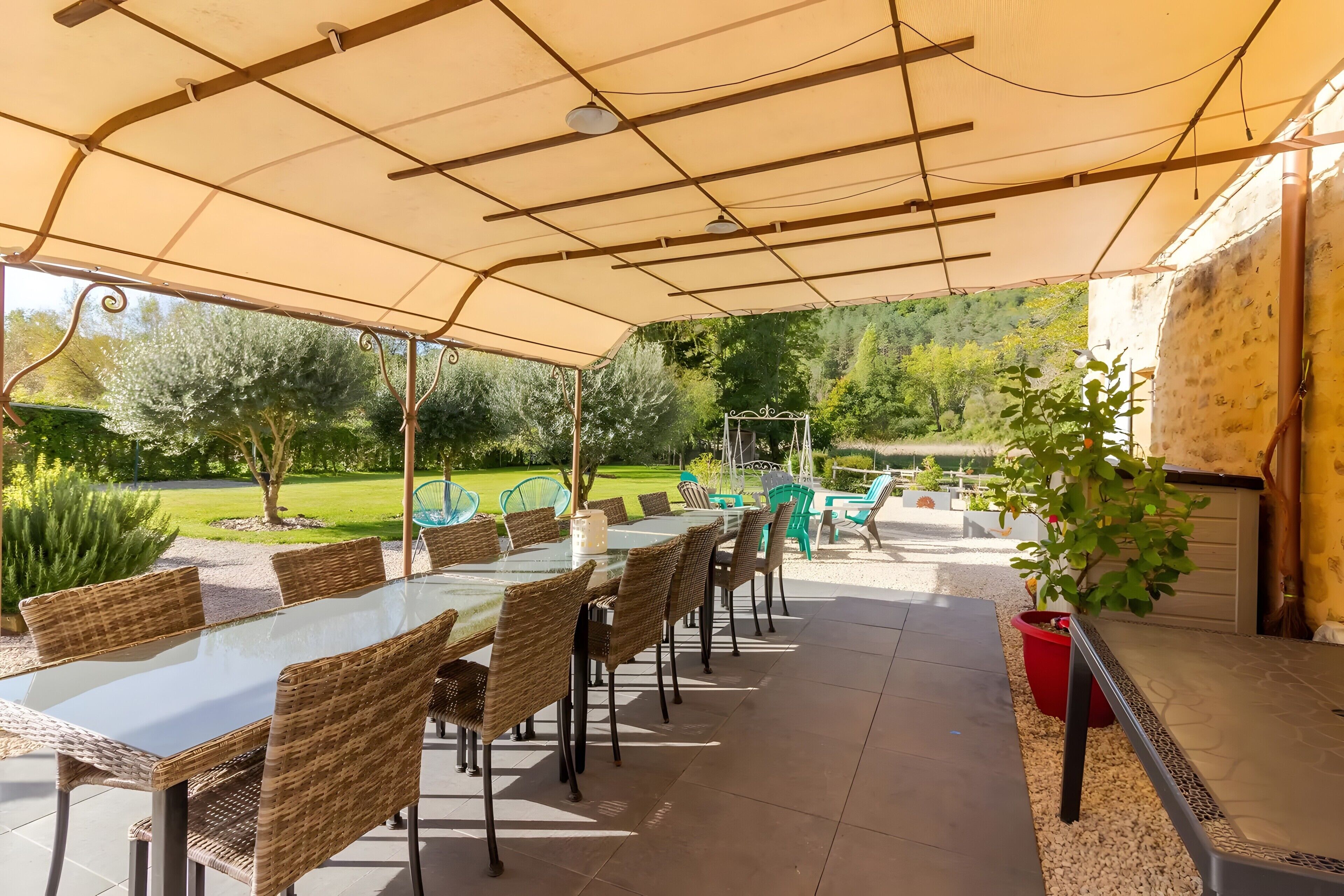 Un patio exterior con una mesa de vidrio y sillas de mimbre, rodeado por una pérgola y vistas a un jardín ajardinado.