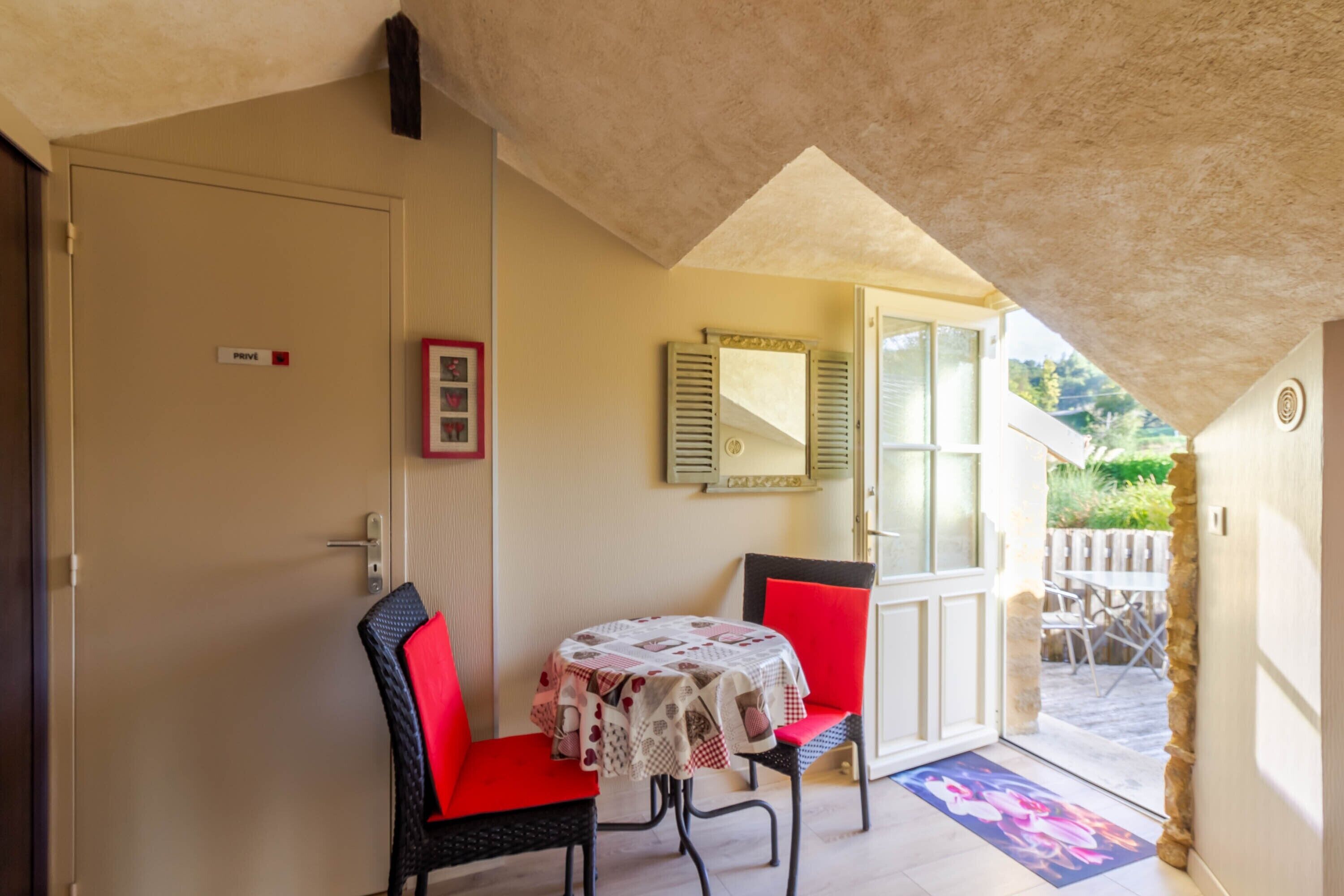 A room with a door marked 'private,' a table with a patterned tablecloth, two red chairs, a mirror, and a view of an outdoor area with a wooden fence and greenery.