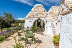 Ein Steinhaus mit einem spitzen Dach, einer gepflasterten Terrasse mit Stühlen und Tischen sowie ein Garten mit violetten Blumen.