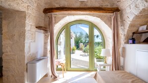 A room with a stone wall, a bed, a radiator, a table with a lamp, and a view through an arched doorway. - Villa 'Trulli E Frasche, Intera' with Private Pool, Wi-Fi and Air Conditioning (Martina Franca)