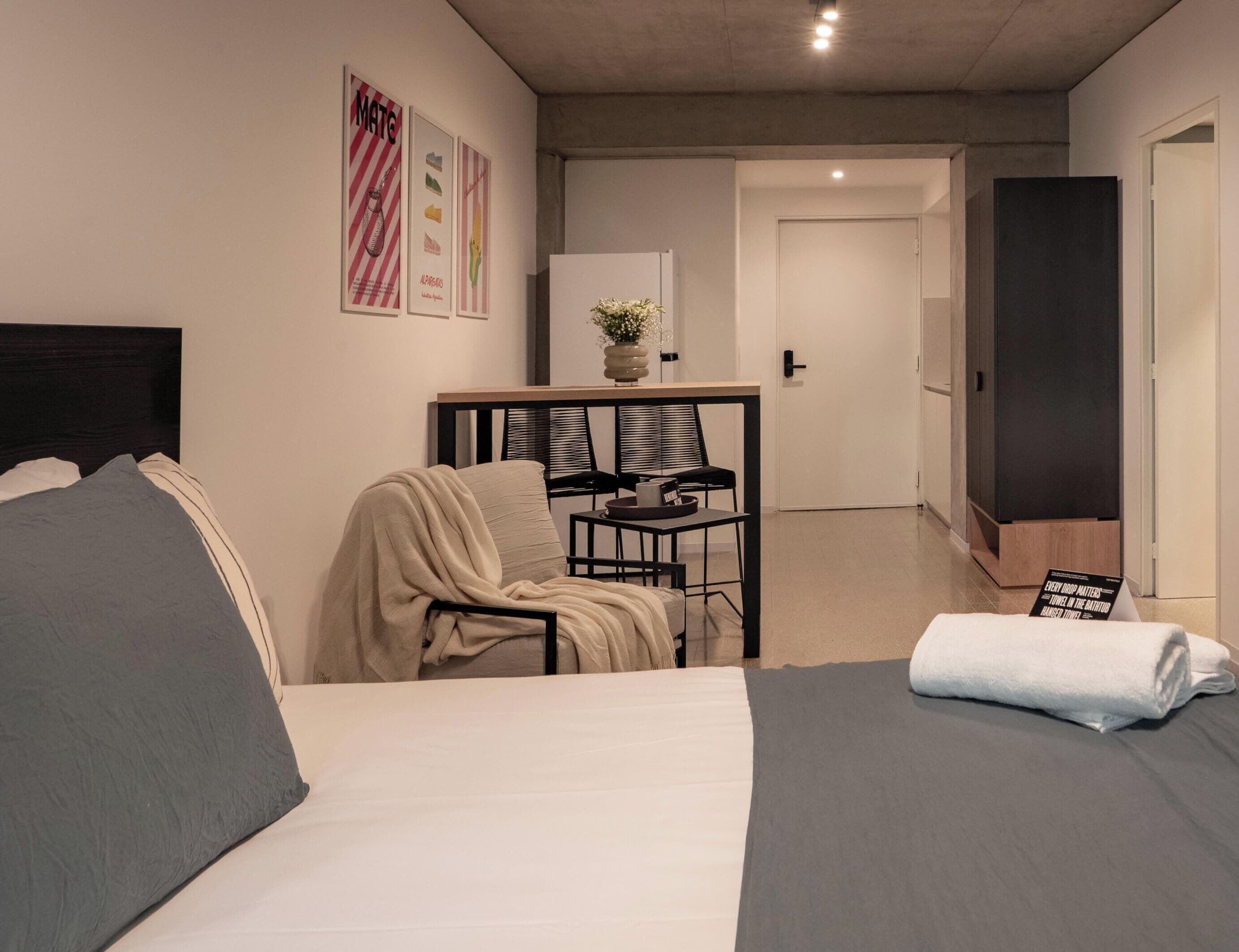 Un dormitorio con dos camas, un escritorio con silla, una mesita con un florero y una puerta que da a otra habitación.