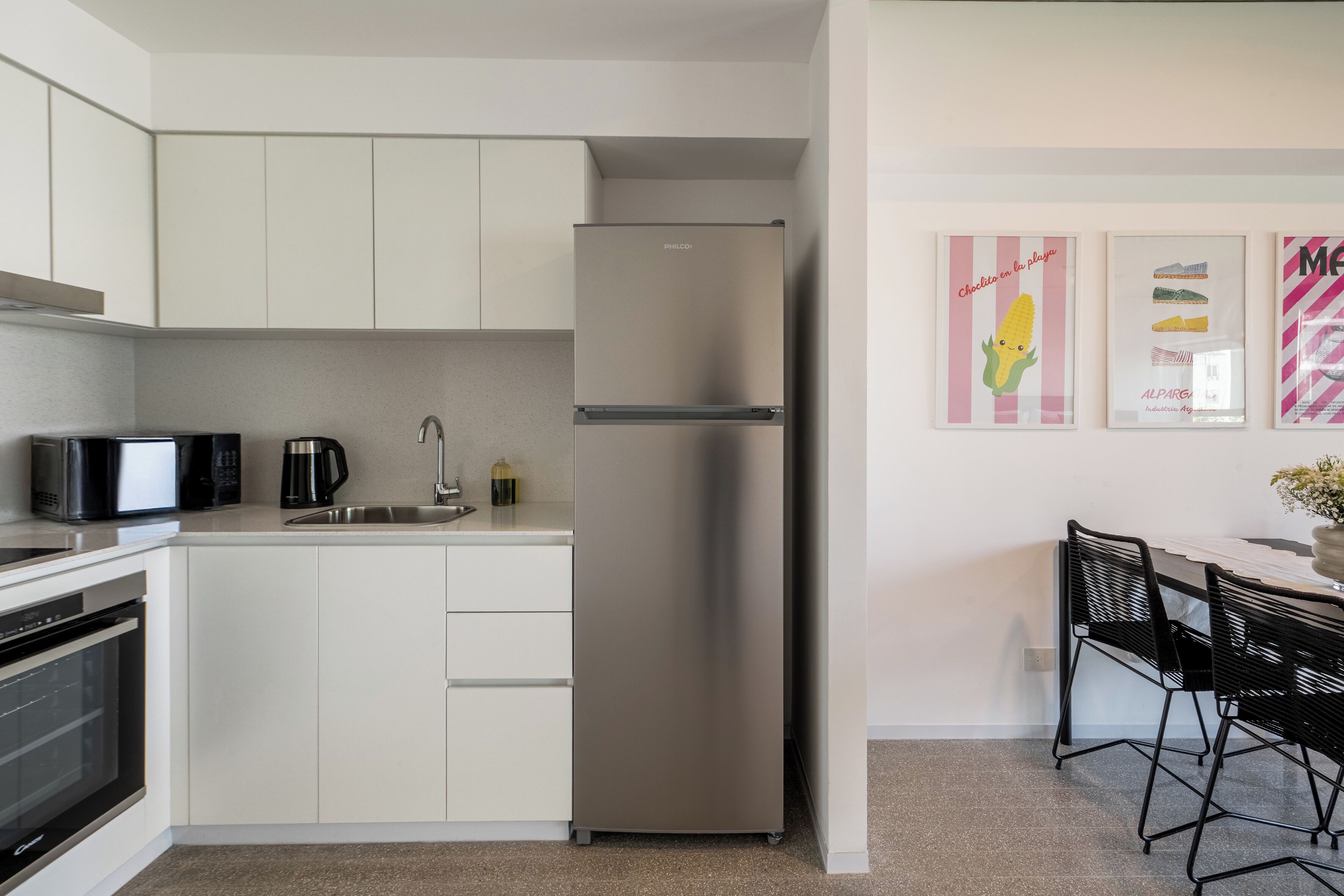 A modern kitchen with white cabinets, a stainless steel refrigerator, a microwave, a coffee maker, and a dining area with black chairs.