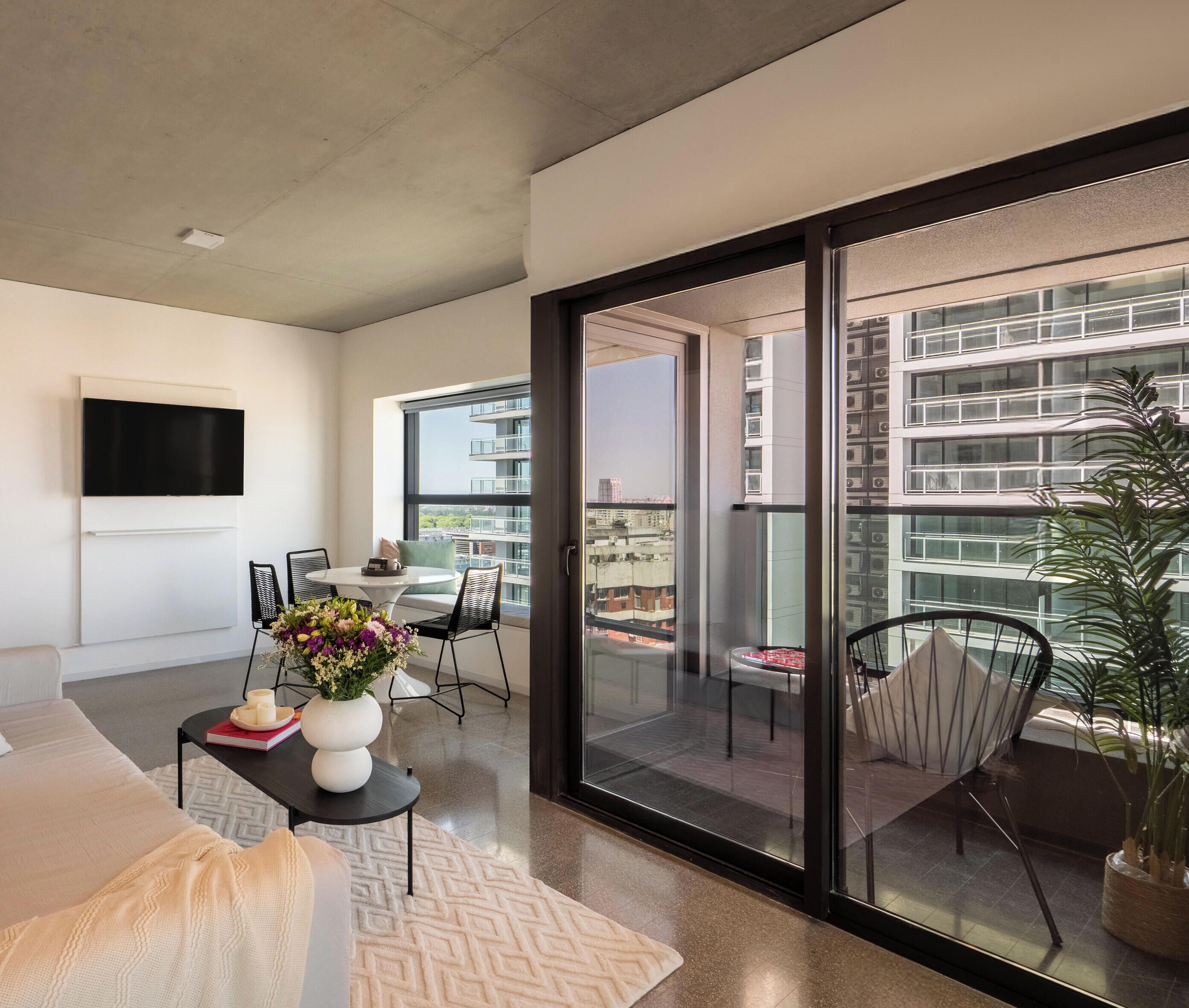 A modern living room with a flat-screen TV, a sofa, a dining table with chairs, a potted plant, and a balcony with a view of the cityscape.